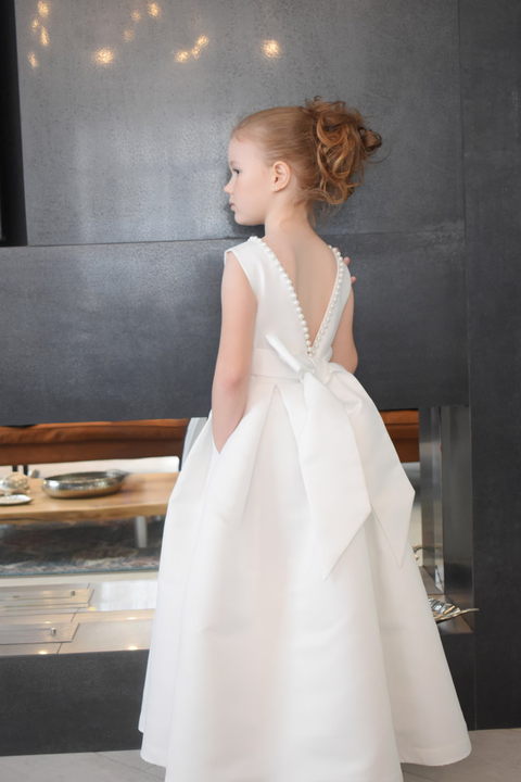Ivory satin flower girl dress with pearl V-back trim, large satin bow and side pocket, dark interior background